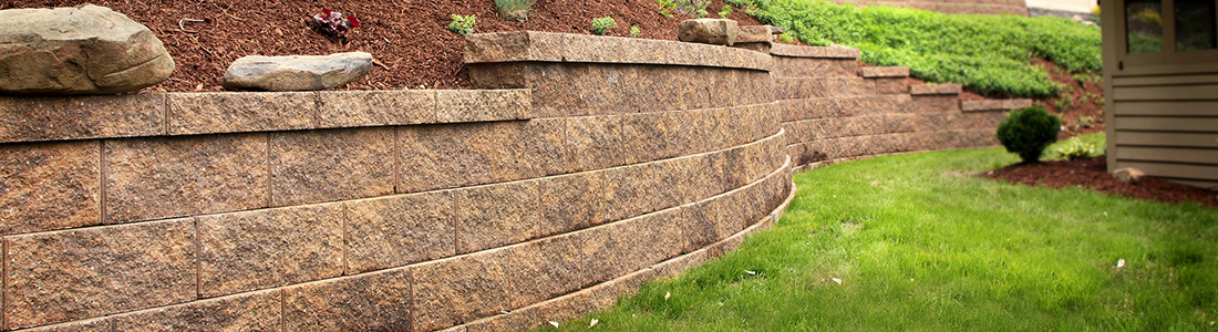 Stone retaining walls with tiered design, enhancing a landscaped garden area with natural stone and lush greenery. Contact Us at The Concrete Service, Inc.